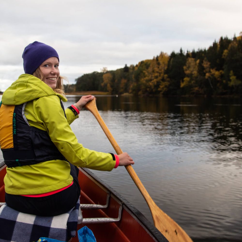 Smiling woman paddling in a canoe, wearing a hat, surrounded by autumn colors in Bogesund Nature Reserve.